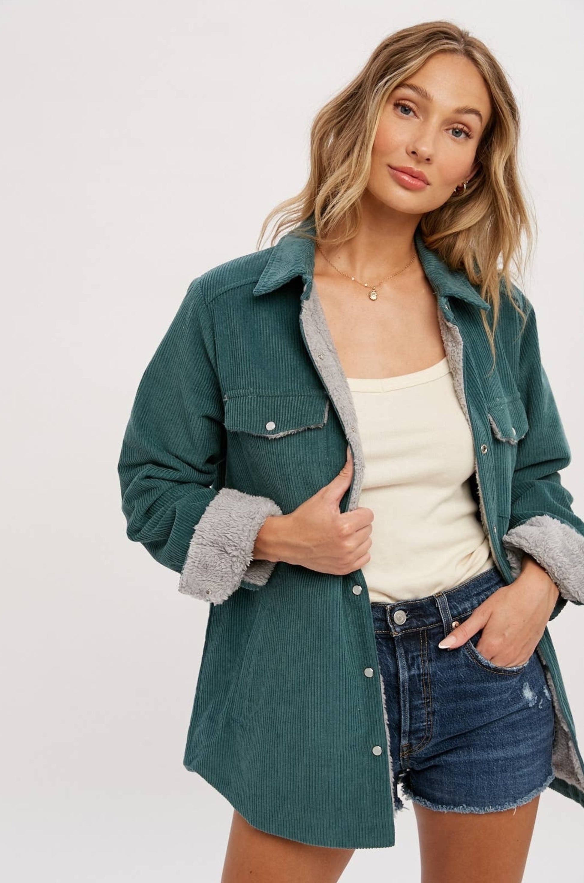 Corduroy jacket featuring a button-down front, long sleeves, sherpa fur lining, and two chest pockets and side pockets. Model has paired outfit with gold necklace, cream undershirt, an distressed dark wash denim shorts. 